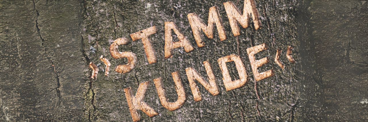 A&J Stammkunde werden und von vielen Vorteilen profitieren – Jetzt Antrag ausfüllen! In der Rinde eines dunklen Baumstamms ist das Wort Stammkunde eingeritzt