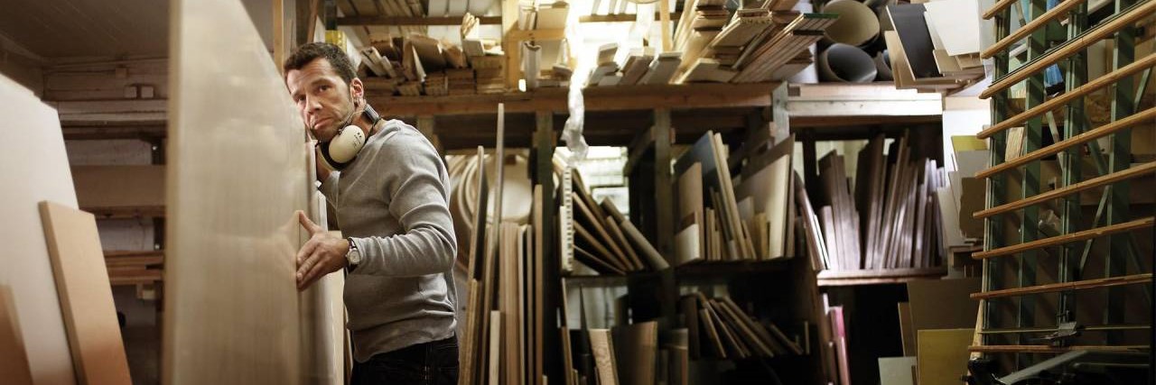 Tischler in einer Holzwerkstatt hantiert mit einer Holzplatte Ein Tischler in einer Holzwerkstatt fässt eine senkrecht stehende Holzplatte an und schaut über den Betrachter hinweg in die Ferne