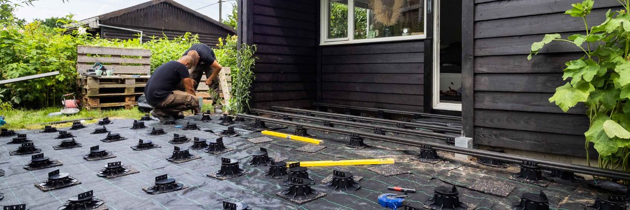 A&J baut WPC Terrasse zusammen mit den Laubengirlz – Anleitung WPC Terrassenbau Zwei Handwerker bauen eine Unterkonstruktion für eine WPC Terrasse auf einer Rasenfläche vor einem schwarzen Gartenhaus